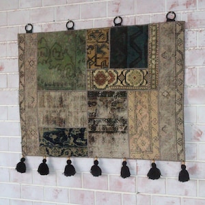 Peut inclure: Une tenture murale rectangulaire compos&eacute;e de divers morceaux de tissu tiss&eacute; dans des tons de vert, marron, beige et noir. La pi&egrave;ce pr&eacute;sente une bordure d&eacute;corative, quatre anneaux m&eacute;talliques noirs en haut pour la suspension et sept pompons noirs.