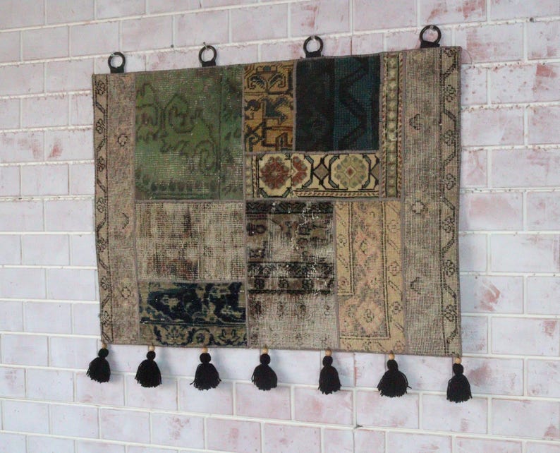 Peut inclure: Une tenture murale rectangulaire compos&eacute;e de divers morceaux de tissu tiss&eacute; dans des tons de vert, marron, beige et noir. La pi&egrave;ce pr&eacute;sente une bordure d&eacute;corative, quatre anneaux m&eacute;talliques noirs en haut pour la suspension et sept pompons noirs.