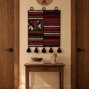 Peut inclure: Une tenture murale tissée rectangulaire présentant des motifs géométriques rouges, noirs et crème, suspendue par trois anneaux métalliques avec cinq pompons bleu foncé en bas. Au-dessus, un plateau panier tissé rond avec un motif en étoile rouge.