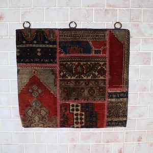 Op de afbeelding: Een rechthoekige patchwork wanddecoratie met verschillende delen van stoffen met patronen. De kleuren zijn rood, bruin, blauw en beige. Het heeft metalen ringen om op te hangen. De afmetingen zijn in centimeters.