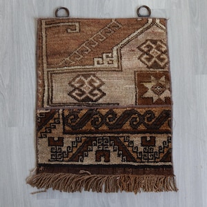 Op de afbeelding: Een rechthoekige, geweven wandhanger met een bruin en beige geometrisch patroon. De bovenkant heeft twee metalen ringhandvatten om op te hangen. De onderste rand heeft een franje. Het ontwerp bevat herhalende vormen en patronen.