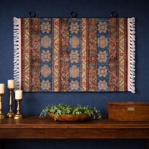 May include: A rectangular wall hanging with a patterned design in shades of blue, brown, and beige, with white fringe. Below, a wooden table holds three gold candle holders with white candles, a wooden bowl with greenery, and a wooden box.