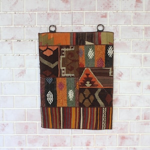 Könnte beinhalten: Ein rechteckiger Wandbehang aus Patchwork mit verschiedenen geometrischen Mustern und Farben. Das Stück weist eine Mischung aus Braun-, Orange-, Grün- und Rottönen auf und hat Metallringe zum Aufhängen. Das Design umfasst Diamanten-, Dreiecks- und Streifenmuster.
