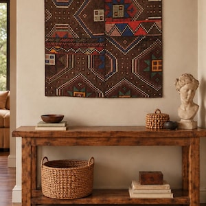 May include: A rectangular brown woven wall hanging featuring geometric patterns in red, blue, and white, suspended by three metal rings. The textile displays intricate diamond and linear motifs across its dark brown surface, creating a structured design.