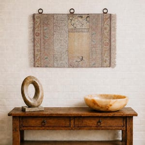 Puede incluir: Una consola de madera rústica con dos cajones, coronada por una escultura de piedra y un cuenco decorativo. Arriba, un tapiz rectangular con un diseño descolorido cuelga contra una pared de ladrillos blancos.