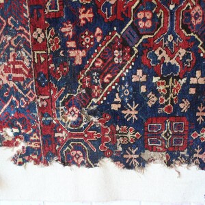 May include: A rectangular woven textile fragment featuring a deep blue and red geometric pattern with floral motifs. The edges are frayed and worn. Two vertical strands of wooden beads and dark blue yarn tassels hang from the top corners.