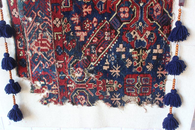 May include: A rectangular woven textile fragment featuring a deep blue and red geometric pattern with floral motifs. The edges are frayed and worn. Two vertical strands of wooden beads and dark blue yarn tassels hang from the top corners.