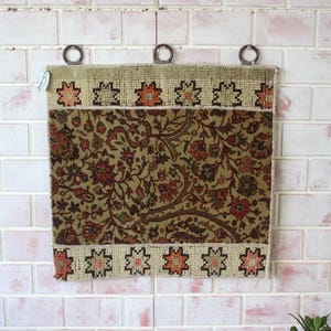 May include: A vintage tapestry wall hanging with a floral pattern in shades of brown, green, and cream. The tapestry has a border with a geometric design in red, white, and black.