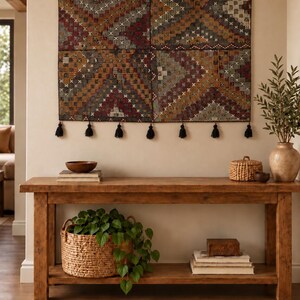 May include: A rectangular wall hanging featuring a multi-colored geometric pattern in shades of red, brown, and grey, accented with small white cross-stitch details. The textile is suspended by six dark metal rings and finished with black tassels.