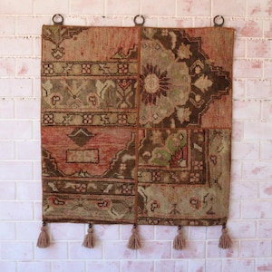 May include: A square wall hanging tapestry featuring a patchwork design of woven fabric in shades of rust, olive green, and dark brown. The textile displays intricate geometric and floral patterns, finished with five hanging tassels along the bottom edge.