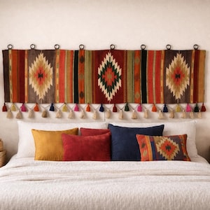 Op de afbeelding: Een slaapkamer met een decoratief hoofdbord, kussens en geweven meubels. Het hoofdbord is een wandtapijt met geometrische patronen in bruin, oranje en groen. Kwasten hangen eronder. Een geweven hanglamp en spiegel maken de inrichting compleet.
