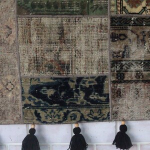 Peut inclure: Un tapis patchwork rectangulaire compos&eacute; de diverses sections tiss&eacute;es dans des tons sourds de beige, marron, vert et noir. Le tapis est constitu&eacute; de panneaux de tissu cousus et fini sur le bord inf&eacute;rieur par cinq pompons en laine noire.