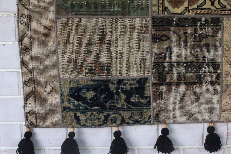 Peut inclure: Un tapis patchwork rectangulaire compos&eacute; de diverses sections tiss&eacute;es dans des tons sourds de beige, marron, vert et noir. Le tapis est constitu&eacute; de panneaux de tissu cousus et fini sur le bord inf&eacute;rieur par cinq pompons en laine noire.
