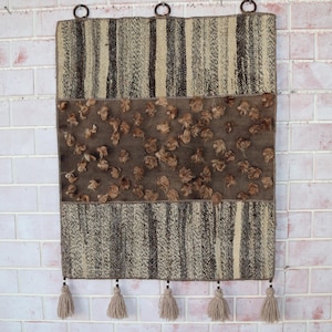 May include: A rectangular wall hanging featuring a woven pattern of dark brown and cream vertical stripes on the top and bottom sections. The center band is a solid dark brown fabric adorned with numerous small, tufted brown fabric rosettes.