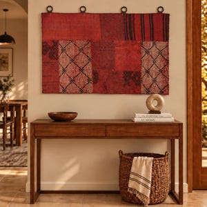 Peut inclure: Une tenture murale rectangulaire composée d'un patchwork de panneaux en tissu rouge avec des motifs géométriques sombres, suspendue par quatre anneaux en métal foncé. Une console en bois porte un bol, une sculpture et deux livres.