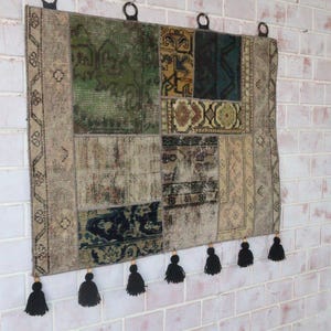 Peut inclure: Une tenture murale rectangulaire compos&eacute;e de segments de tapis en patchwork dans des tons de vert, bleu, beige et marron. Le textile pr&eacute;sente divers motifs g&eacute;om&eacute;triques et floraux, trois anneaux de suspension noirs et six pompons noirs.