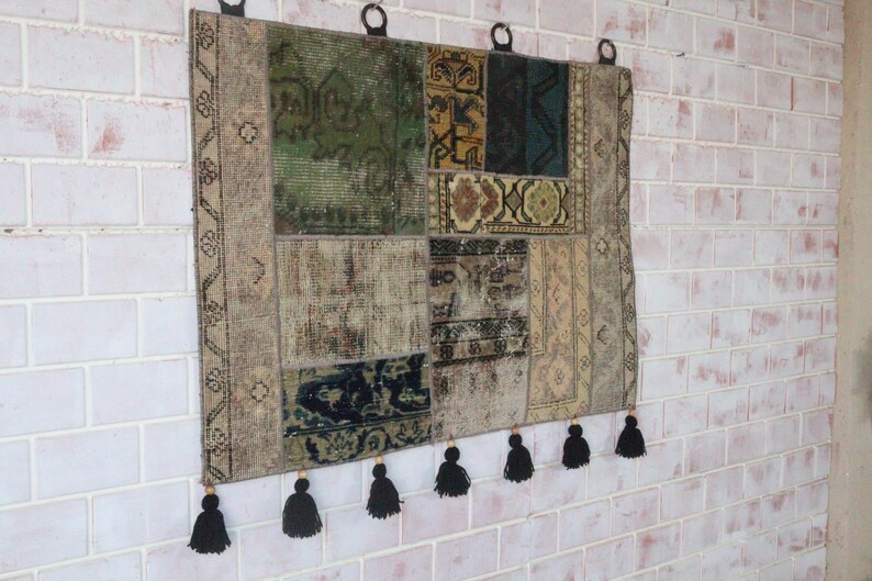 Peut inclure: Une tenture murale rectangulaire compos&eacute;e de segments de tapis en patchwork dans des tons de vert, bleu, beige et marron. Le textile pr&eacute;sente divers motifs g&eacute;om&eacute;triques et floraux, trois anneaux de suspension noirs et six pompons noirs.