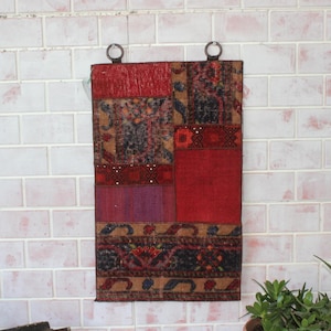May include: A rectangular patchwork wall hanging featuring a mix of red, brown, and purple patterned fabrics. It has two metal rings for hanging. The design incorporates geometric and floral patterns, giving it a rustic, vintage appearance.