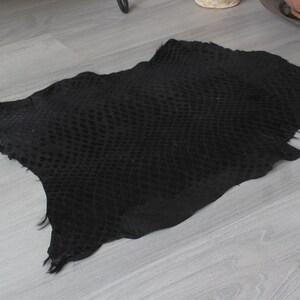 May include: A black snakeskin hide with a textured pattern. The hide is laid flat on a light gray wood floor.