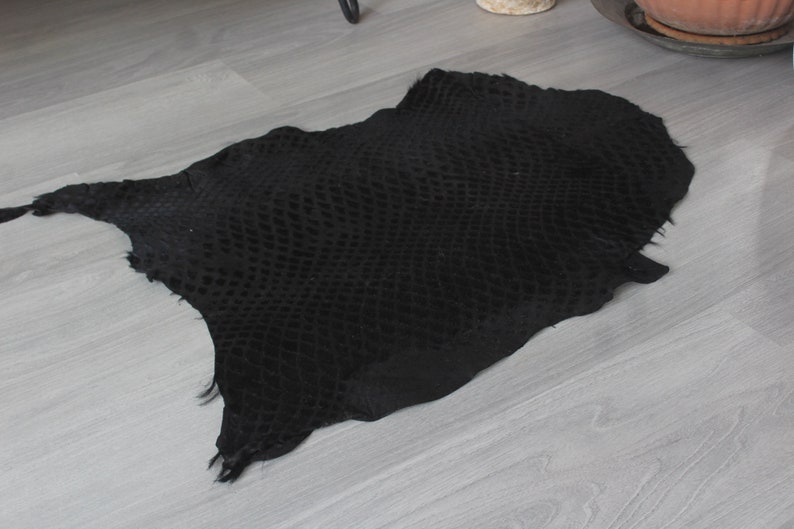 May include: A black snakeskin hide with a textured pattern. The hide is laid flat on a light gray wood floor.
