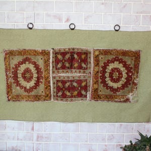 Puede incluir: Un tapiz verde con tres paneles bordados con motivos geométricos rojos, blancos y dorados. Los paneles están unidos al tapiz con anillos de metal negro.