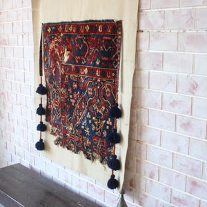 May include: A rectangular wall hanging featuring a dark blue and red patterned textile mounted on a cream-coloured fabric backing. The textile has frayed bottom edges and is decorated with vertical rows of dark blue tassels on both sides.