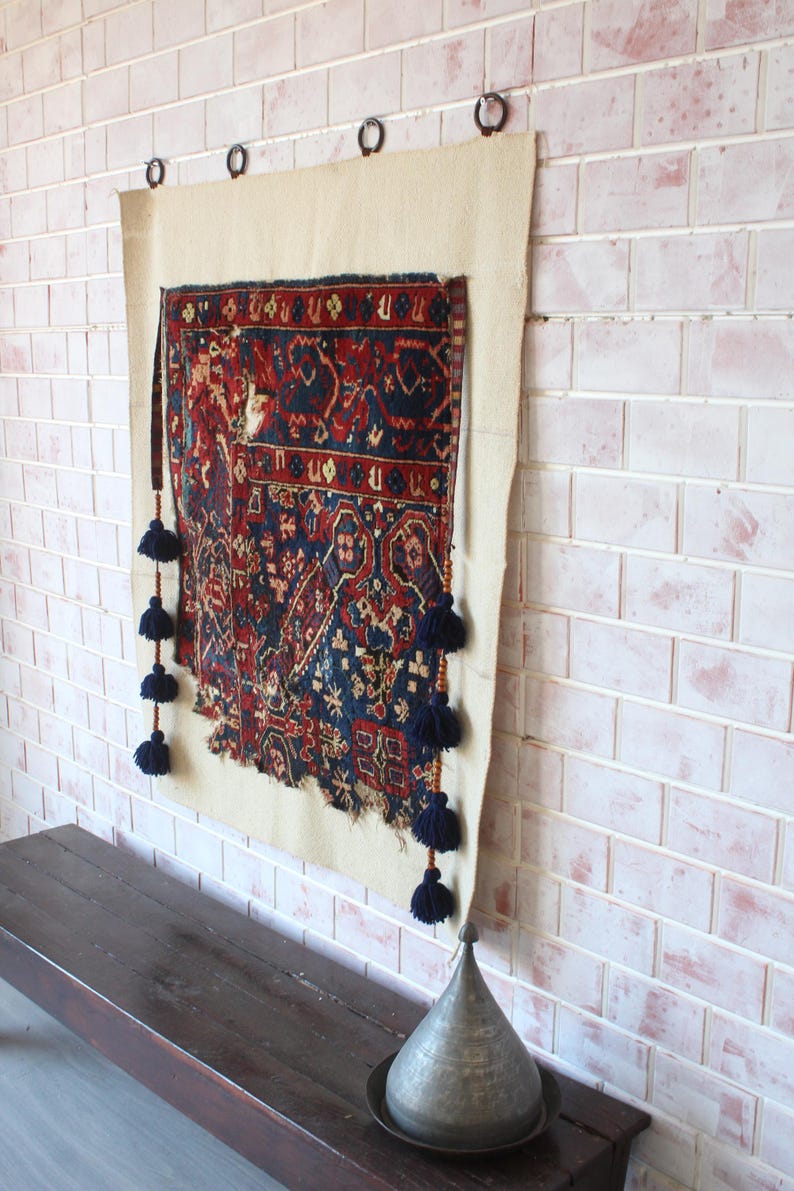 May include: A rectangular wall hanging featuring a dark blue and red patterned textile mounted on a cream-coloured fabric backing. The textile has frayed bottom edges and is decorated with vertical rows of dark blue tassels on both sides.