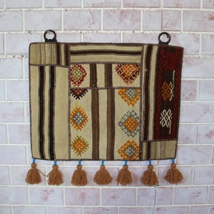 Op de afbeelding: Rechthoekige geweven wandkleed met een patchwork-ontwerp. Het wandtapijt heeft verticale strepen, geometrische patronen en een rijk kleurenpalet van beige, bruin en rood. Bruine kwasten hangen aan de onderkant en metalen ringen aan de bovenkant om op te hangen.