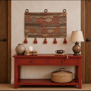 May include: A rectangular wall hanging featuring a geometric pattern in shades of rust, brown, and cream, suspended by three dark rings with five hanging tassels. The textile is displayed above a red wooden console table with two drawers.