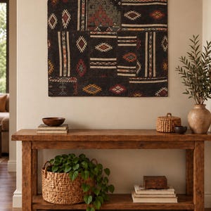 Könnte beinhalten: Ein rechteckiger Wandbehang aus einem dunkelbraunen Patchwork-Textil mit geometrischen Mustern in Weiß, Rot und Gelb. Das Stück ist an vier kreisförmigen Metallringen an der Oberkante befestigt und weist eine Vielzahl von gewebten Texturen auf.