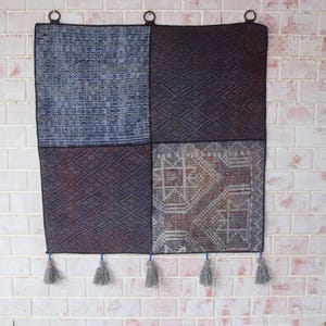 Op de afbeelding: Een vierkante wandhanger met een patchworkontwerp van vier panelen met variërende geometrische patronen in tinten blauw, bruin en grijs. Het textielstuk bevat drie cirkelvormige metalen ringen aan de bovenkant en vijf grijze kwastjes aan de onderkant.
