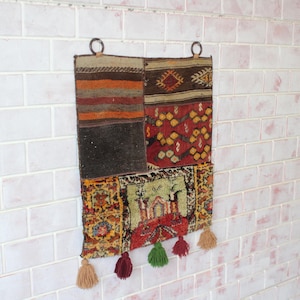 Peut inclure: Une tapisserie murale en tissu patchwork coloré avec des glands. Le tissu est un mélange de rouge, orange, jaune, vert, bleu et marron. La tapisserie est fixée au mur avec deux anneaux métalliques.