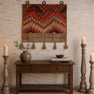 May include: A rustic interior scene featuring a woven wall hanging with geometric patterns in red, orange, and beige tones. A wooden console table holds a vase, books, and a bowl. Tall wooden candle holders with lit candles add warmth.
