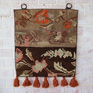 Peut inclure: Une tenture murale rectangulaire tissée avec un motif floral dans les tons de marron, orange et rose. Le haut comporte deux anneaux en bois pour la suspension. Le bord inférieur comporte six pompons orange.