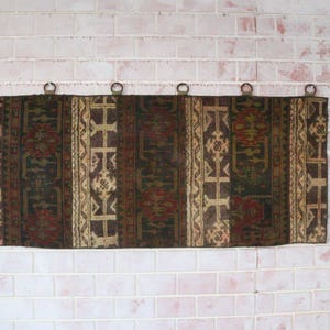 Puede incluir: Un tapiz rectangular con una serie de paneles verticales con intrincados patrones geométricos y florales en tonos rojo oscuro, marrón, negro y crema. El tejido está suspendido de seis anillos metálicos circulares en el borde superior.