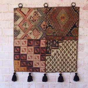 Op de afbeelding: Een vierkante wandhanger met een patchworkontwerp van vier verschillende geweven textielpanelen in tinten rood, bruin, crème en zwart. Het stuk bevat geometrische patronen, drie metalen ophangringen aan de bovenkant en vijf zwarte kwastjes.