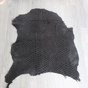 May include: A black snakeskin hide with a textured pattern. The hide is laid flat on a light wood floor.