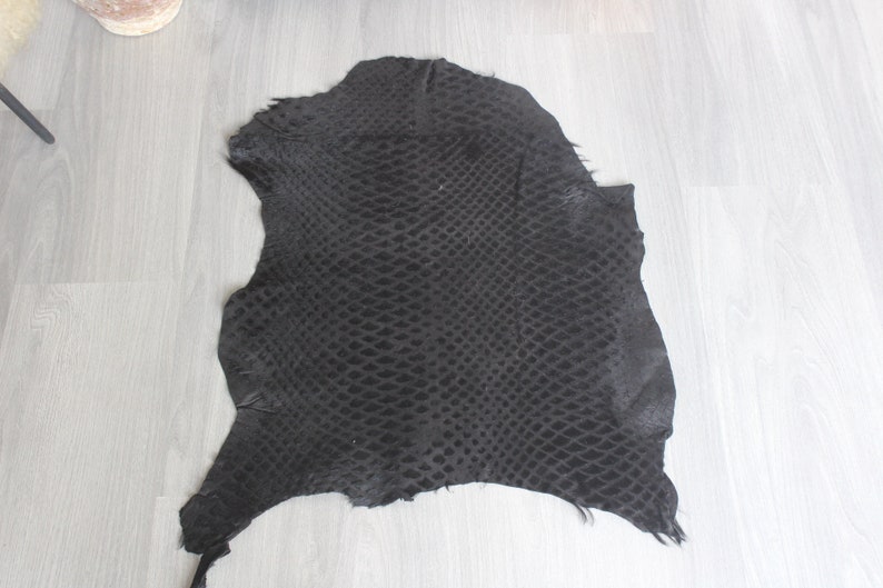 May include: A black snakeskin hide with a textured pattern. The hide is laid flat on a light wood floor.
