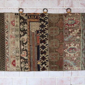 May include: A rectangular wall hanging made of multiple vertical strips of woven fabric featuring diverse patterns in shades of brown, beige, black, and red. The piece includes five circular metal rings along the top edge and white fringe.