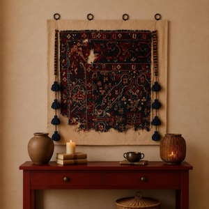 May include: A rectangular wall hanging featuring a dark red and navy blue patterned textile fragment mounted on a beige fabric backing. The piece is accented with vertical side borders, decorative beads, and three navy blue tassels on each side.