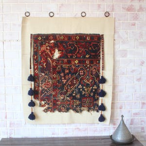 May include: A rectangular wall hanging featuring a distressed, dark-toned woven textile with intricate red, blue, and black geometric patterns. The textile is mounted on a light beige fabric backing and decorated with dark blue tassels on both sides.