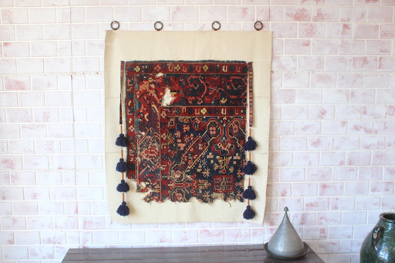 May include: A rectangular wall hanging featuring a distressed, dark-toned woven textile with intricate red, blue, and black geometric patterns. The textile is mounted on a light beige fabric backing and decorated with dark blue tassels on both sides.