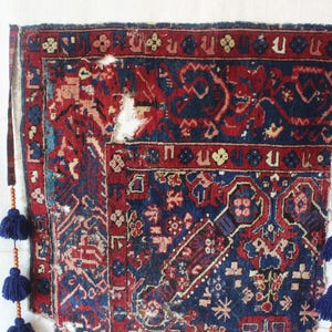 May include: A rectangular woven rug featuring a deep blue and red geometric pattern with visible wear and fraying. The rug is suspended by two vertical fabric straps adorned with wooden beads and dark blue tassels hanging from each side.