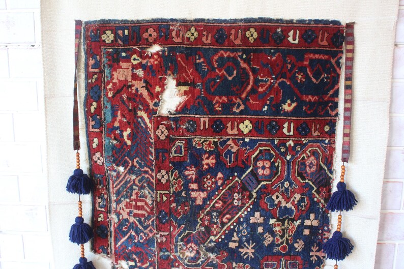 May include: A rectangular woven rug featuring a deep blue and red geometric pattern with visible wear and fraying. The rug is suspended by two vertical fabric straps adorned with wooden beads and dark blue tassels hanging from each side.