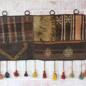 Pode incluir: Uma tapeçaria de parede retangular em patchwork com uma variedade de tecidos castanhos e bege. A parte superior tem quatro anéis de metal para pendurar. A borda inferior apresenta borlas coloridas em tons de vermelho, amarelo e azul.