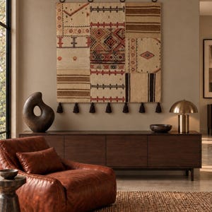 Peut inclure: Une tapisserie murale rectangulaire composée de panneaux de tissu tissé en patchwork aux tons crème, marron et rouge avec des motifs géométriques. L'ensemble est suspendu par quatre anneaux en métal foncé et orné d'une rangée de pompons marron.