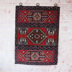 May include: A rectangular wall hanging featuring a red, black, and brown geometric pattern. The textile is divided into three horizontal sections with intricate diamond and border motifs, and it includes three metal rings at the top for hanging.