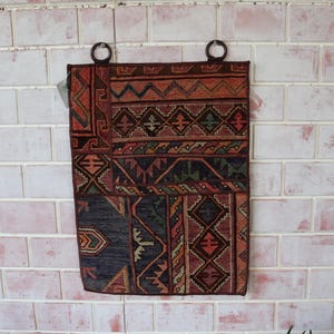 May include: A colorful tapestry with geometric patterns in shades of red, orange, brown, blue, and green. The tapestry is hanging on a brick wall with two metal rings at the top.