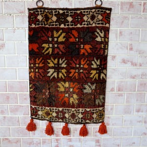 Peut inclure: Une tenture murale rectangulaire tissée avec un motif géométrique dans les tons de marron, rouge, orange et beige. Le motif présente des motifs floraux et géométriques répétés. Le bord inférieur comporte des pompons orange et le haut des anneaux en bois pour l'accrocher.