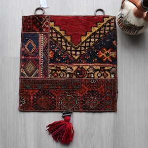 Puede incluir: Una alfombra cuadrada de patchwork con una combinación de colores rojo, azul marino y dorado. La alfombra presenta patrones geométricos y una borla roja. Dos anillos metálicos están unidos en la parte superior para colgar.